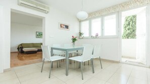 A bright living room with a glass-top dining table surrounded by white chairs, a green sofa, and a window with a view of greenery.