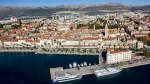 Vista aérea de una ciudad costera con un paseo marítimo destacado, un edificio grande de color blanco con techo rojo, y un crucero atracado en el puerto.