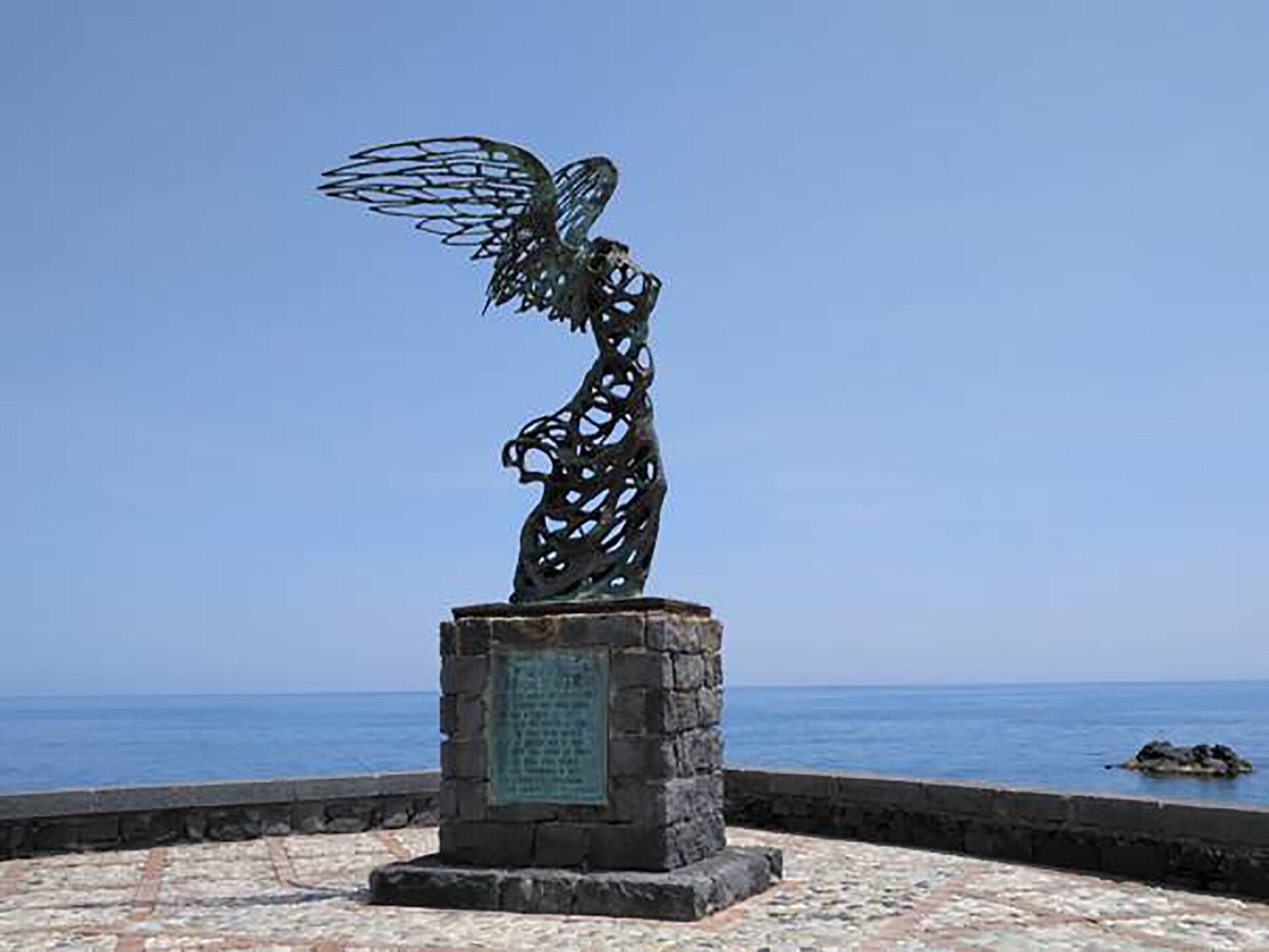 A metal sculpture of an angel on a stone pedestal by the sea.