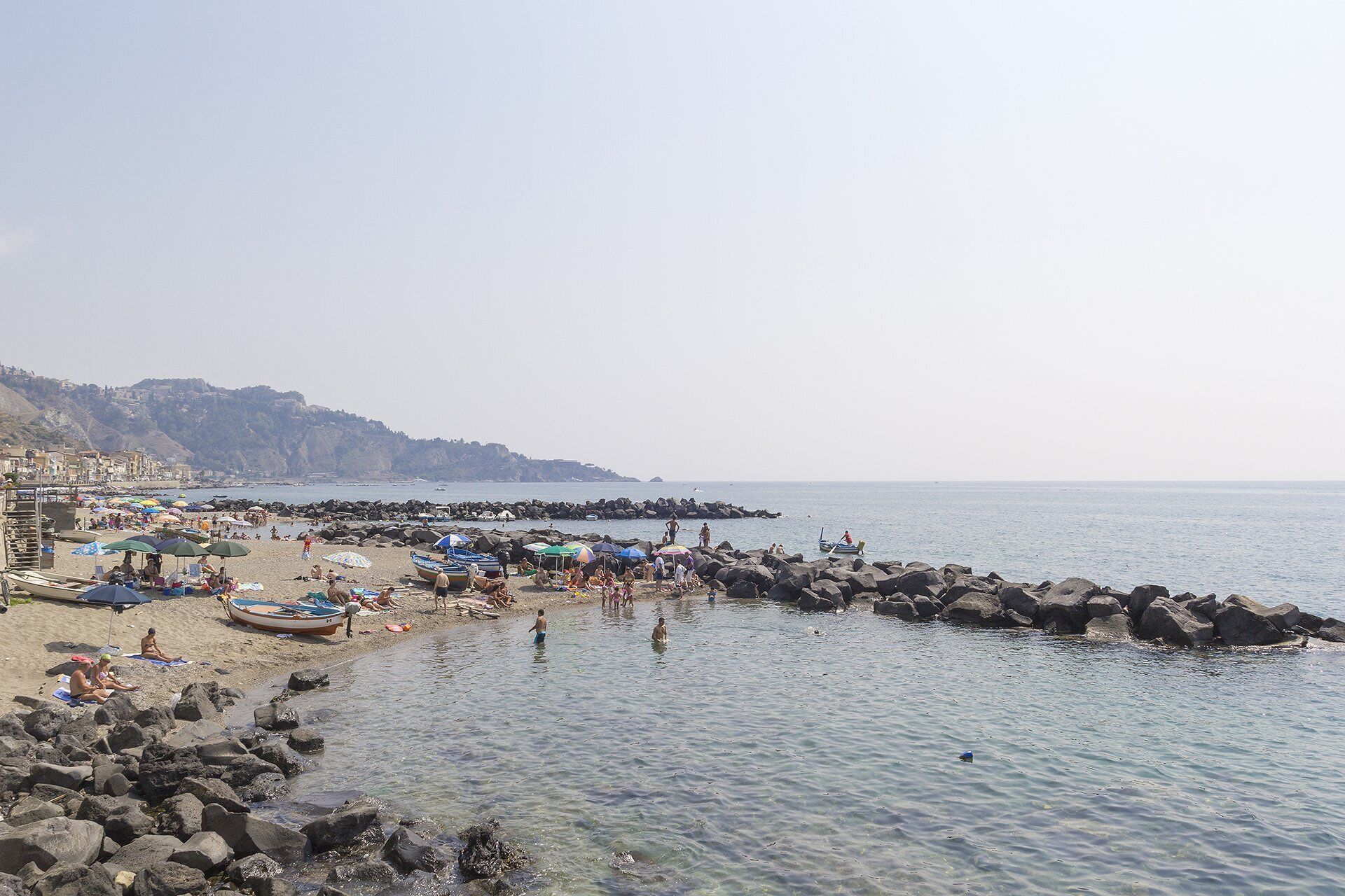 A beach with people sunbathing and swimming, rocky shore, and boats.