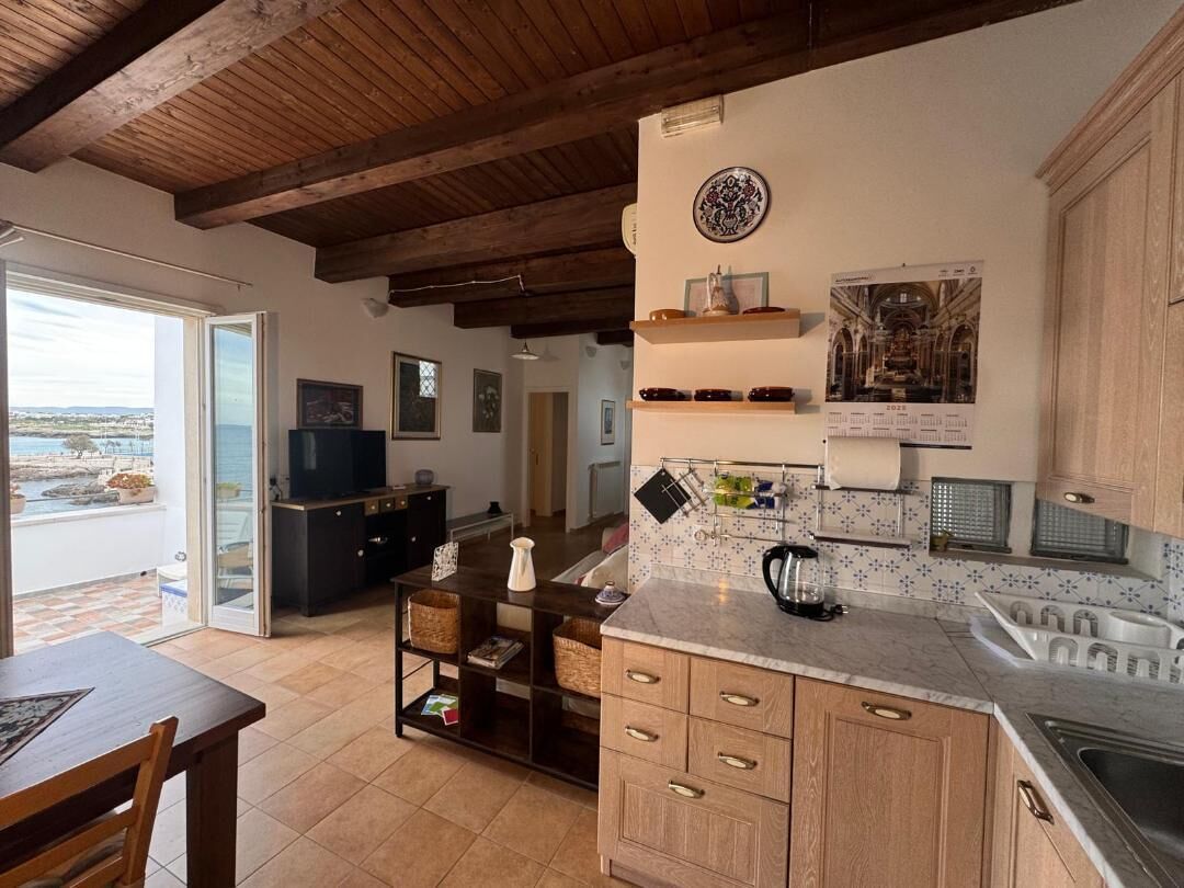 Une cuisine avec des meubles en bois, un plan de travail en marbre et une vue sur la mer, visible à travers une porte ouverte.