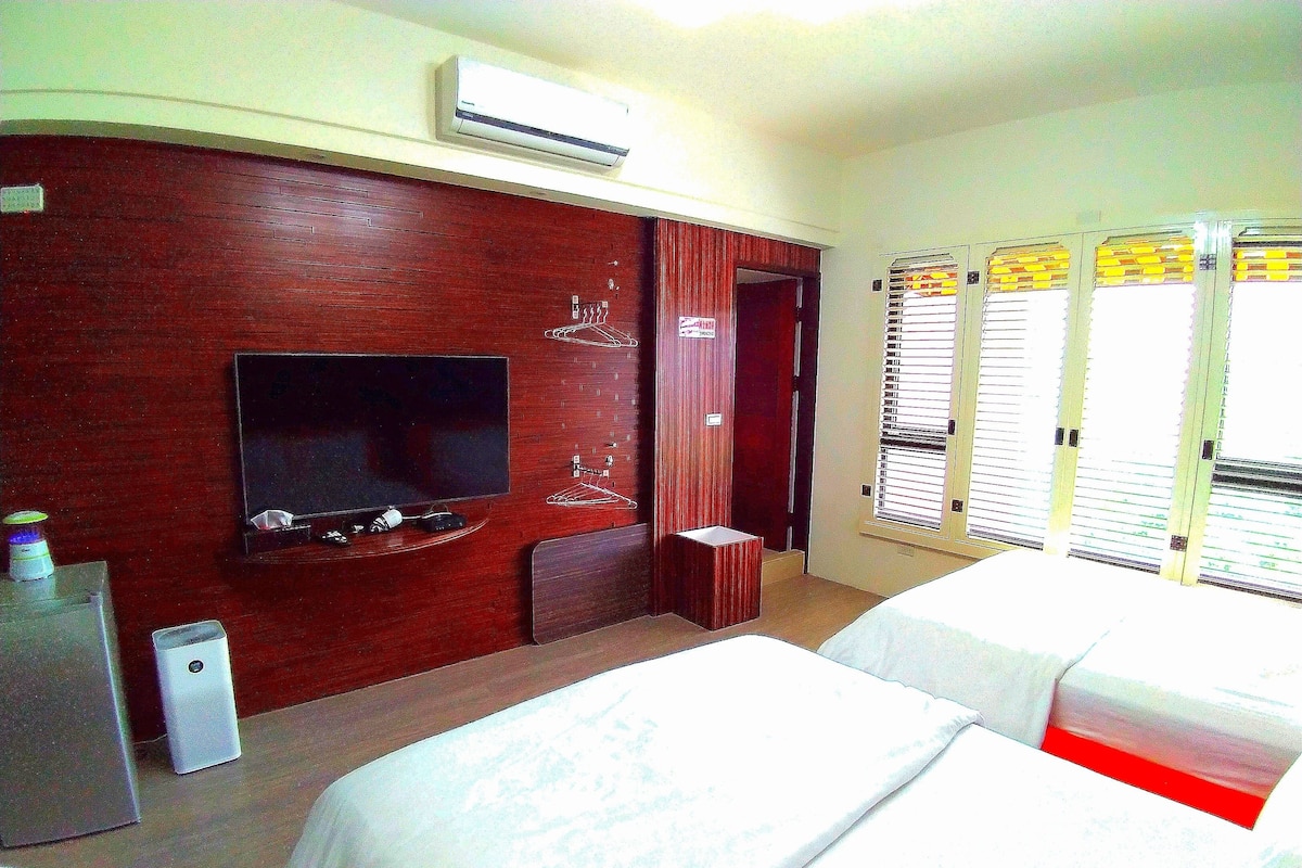 A hotel room with a flat-screen TV, a wooden panel wall, a window with blinds, and a bed with white bedding.