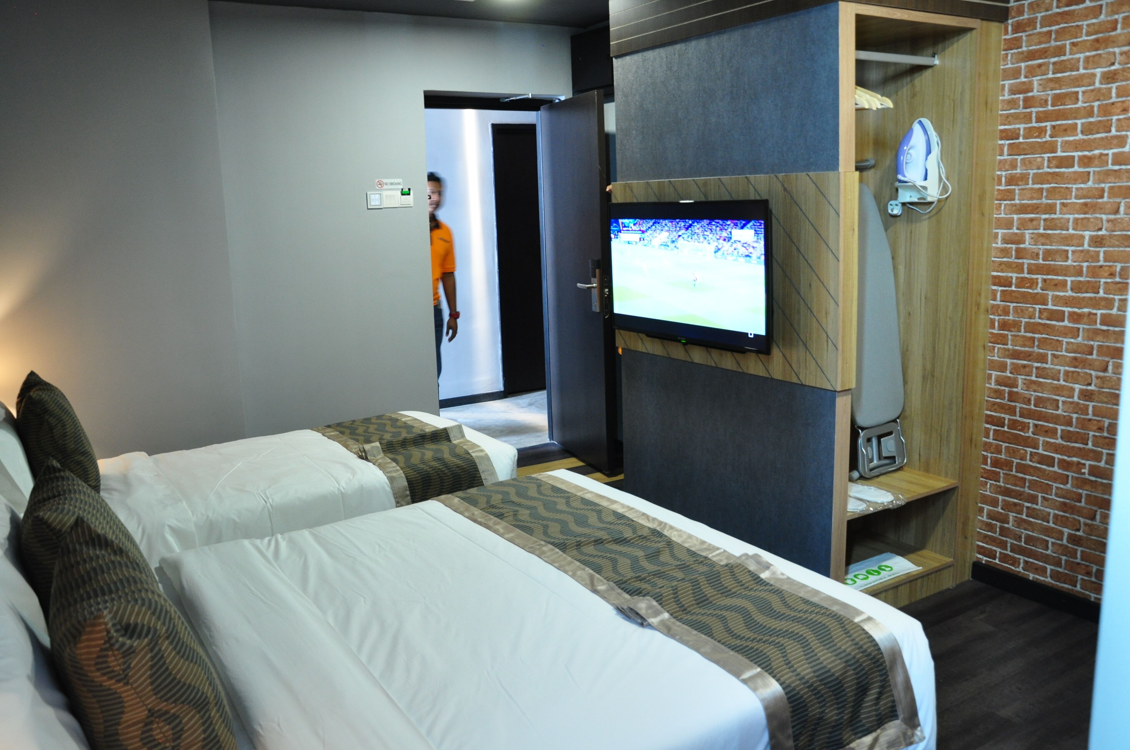 A hotel room with two beds, a TV mounted on the wall, a shower area, and a brick wall.