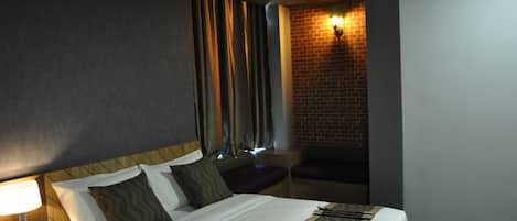 A hotel room with a large bed, a nightstand with a lamp, a brick wall, and a window with curtains.