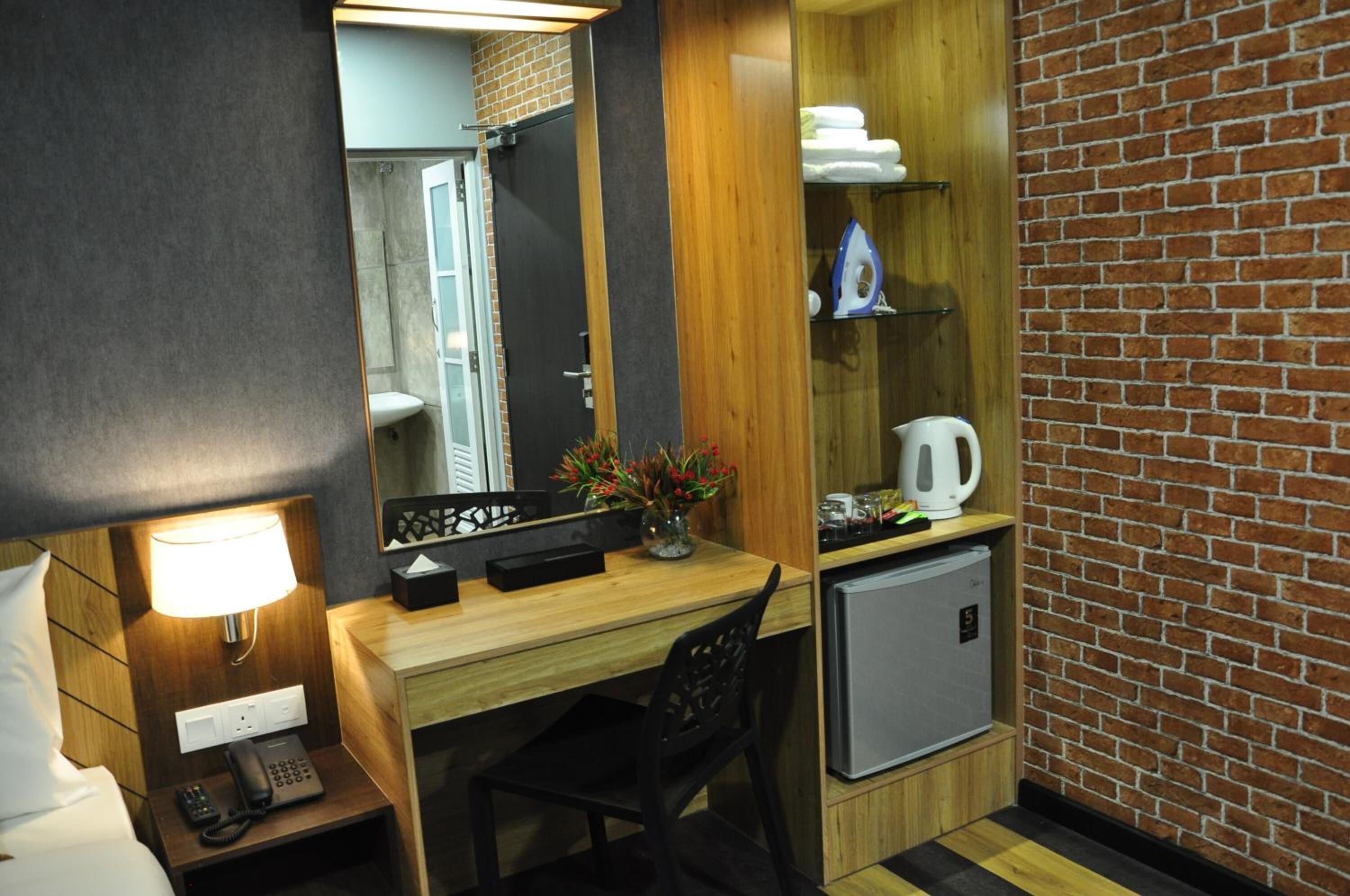 A compact hotel room with a wooden desk, a small refrigerator, a bedside table with a lamp, a telephone, and a mirror.