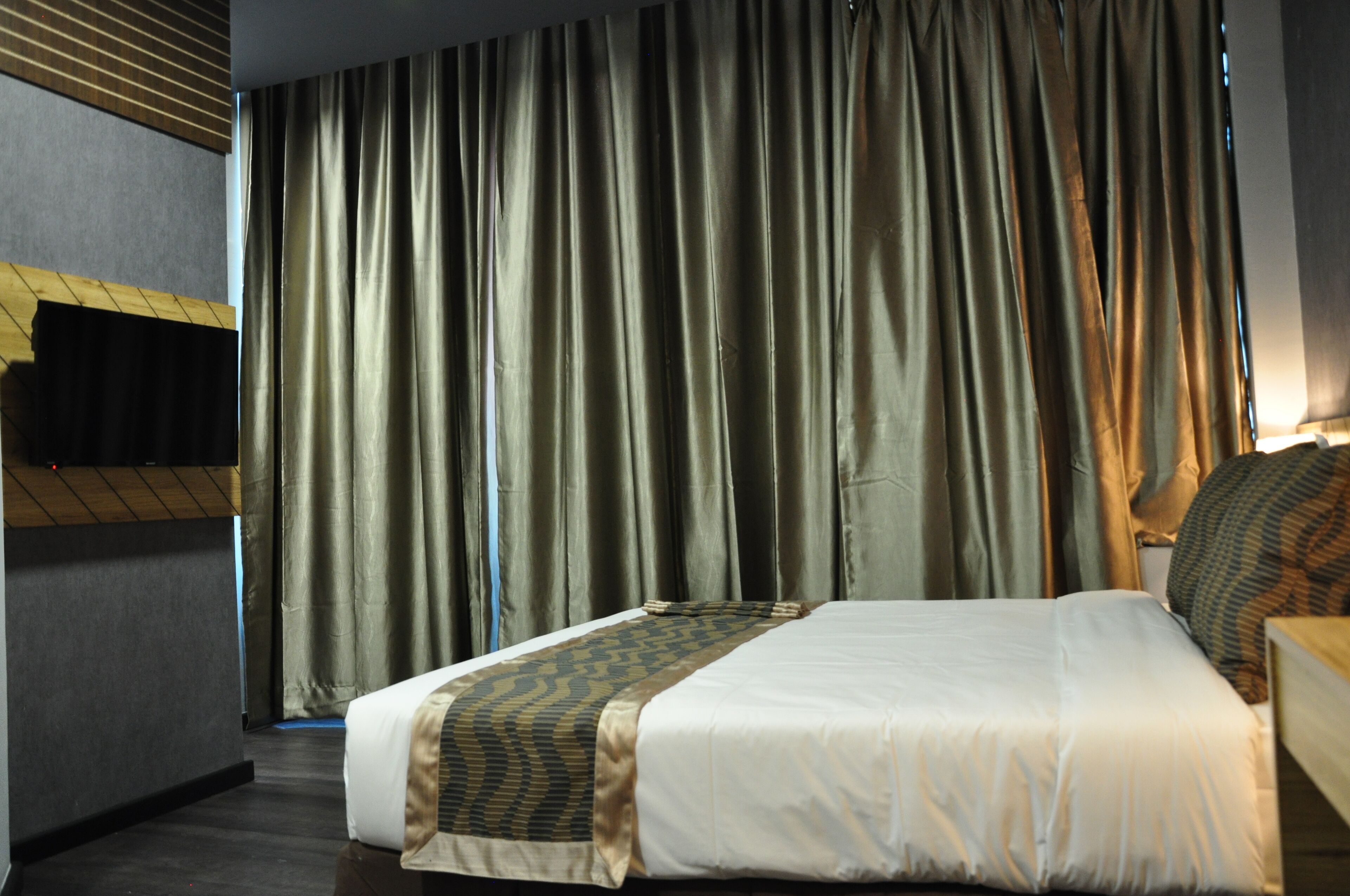 Une chambre d’hôtel avec un lit, des rideaux, un téléviseur fixé au mur et une tête de lit en bois.