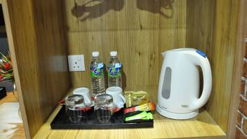 A wooden shelf with a white electric kettle, two bottles of water, a tray with glasses and tea bags, and a power outlet.