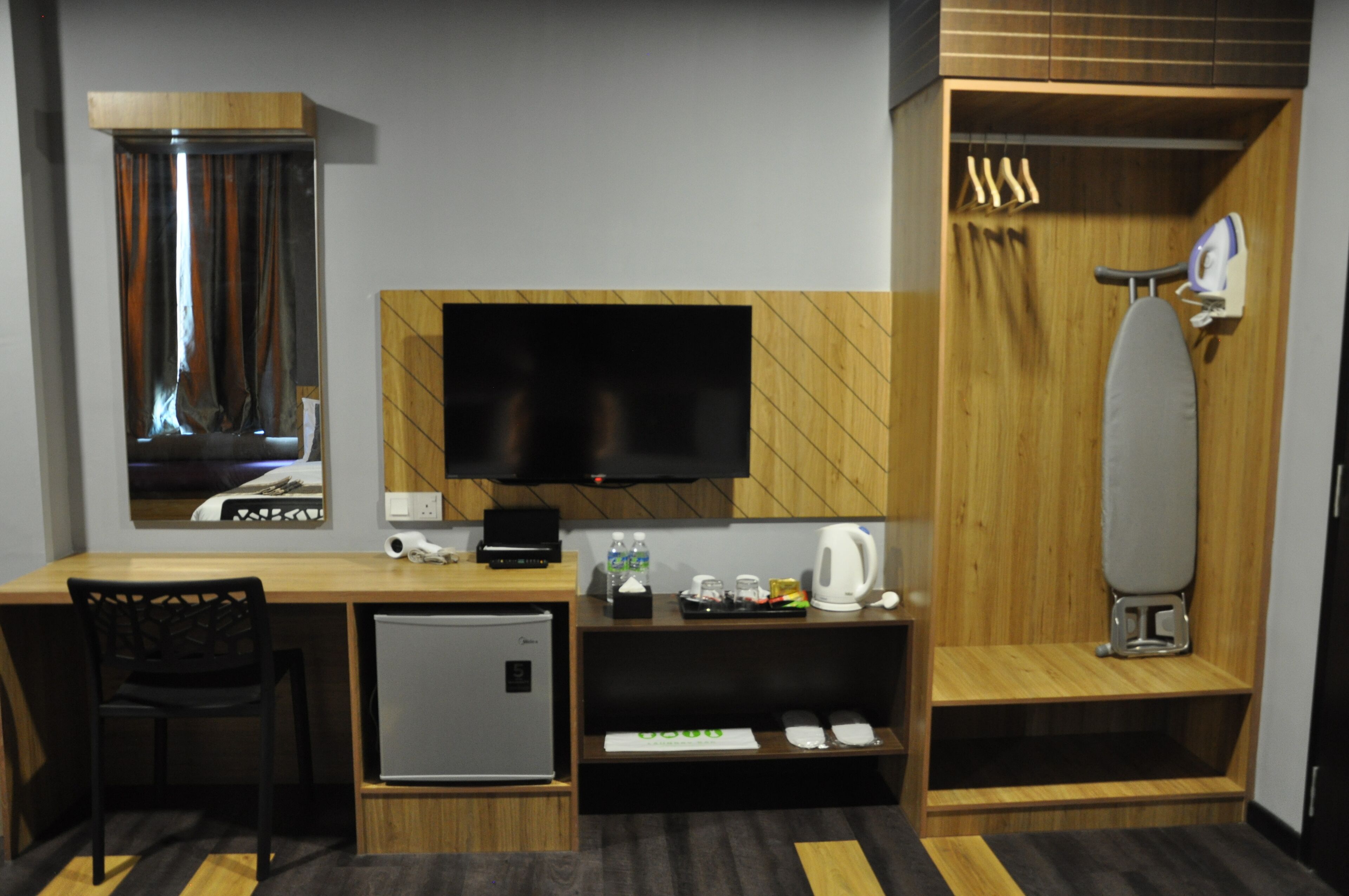 A compact hotel room with a wooden desk, a small refrigerator, a television mounted on the wall, a mirror, a shower area with a towel rack, and a small shelf with toiletries.