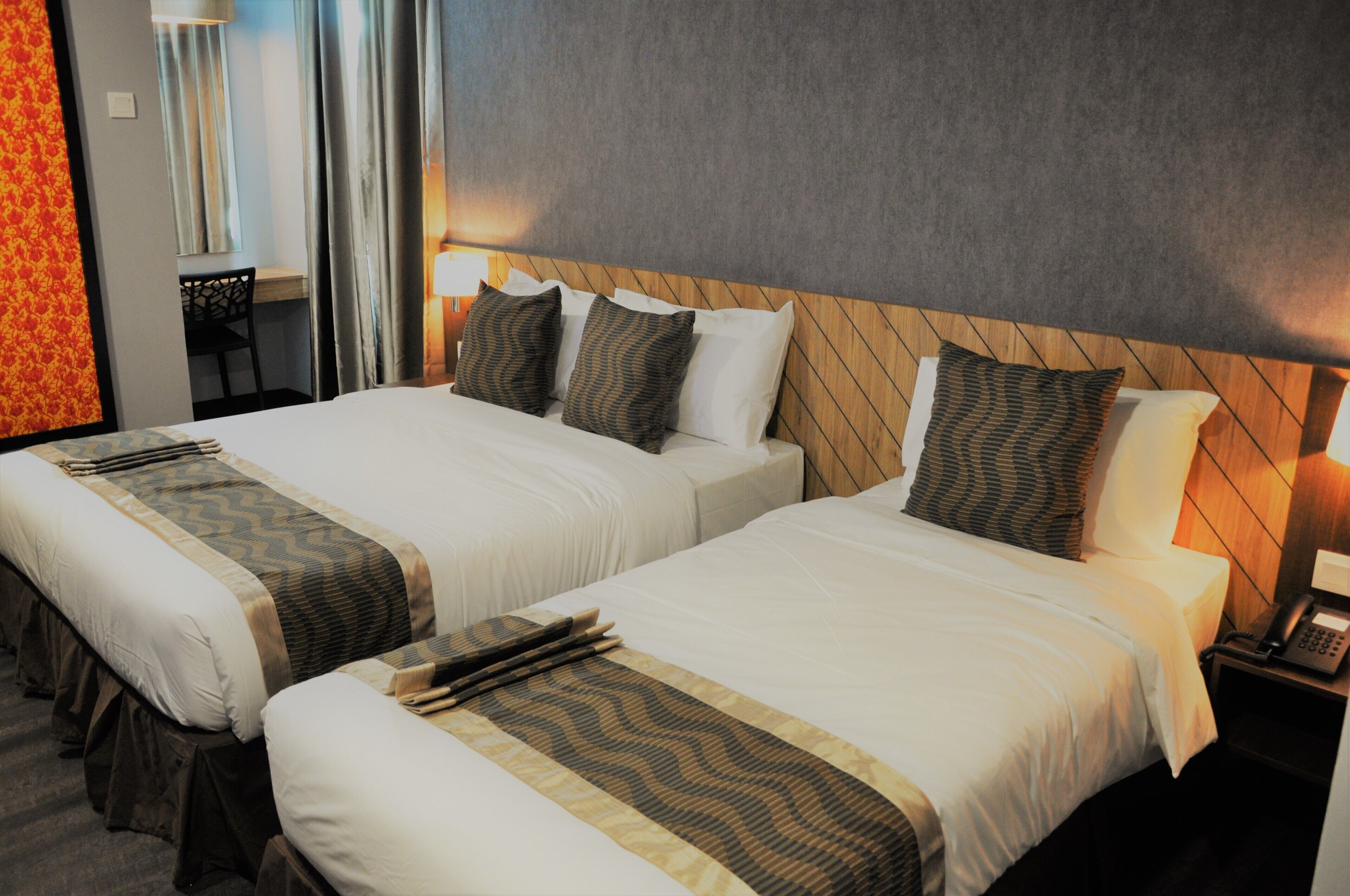 A hotel room with two beds, each with a wooden headboard, white bedding, and patterned throw pillows. There is a desk with a chair and a lamp in the corner.