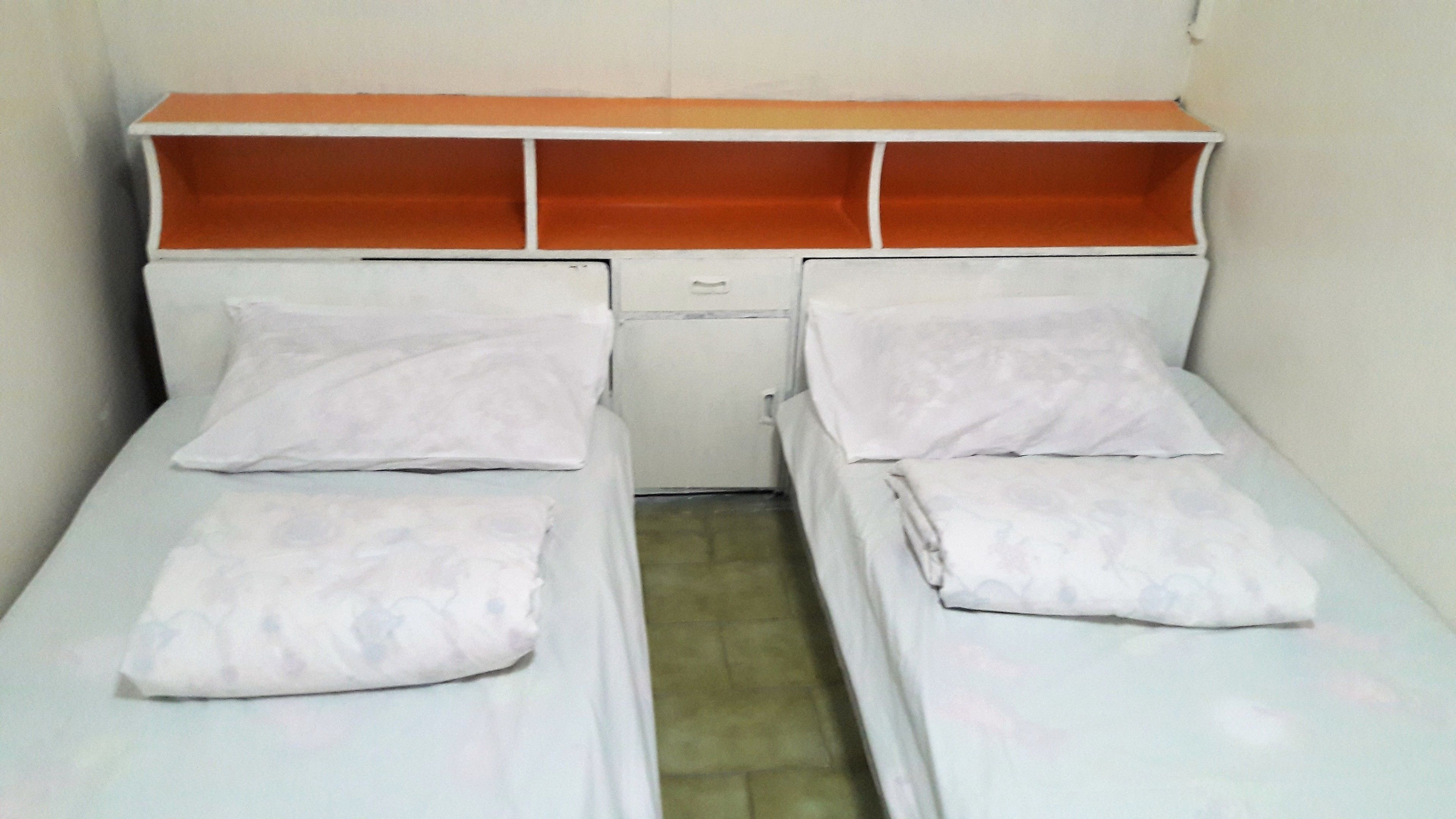 A small, single-room with two beds, a small table, and a shelf above the beds.