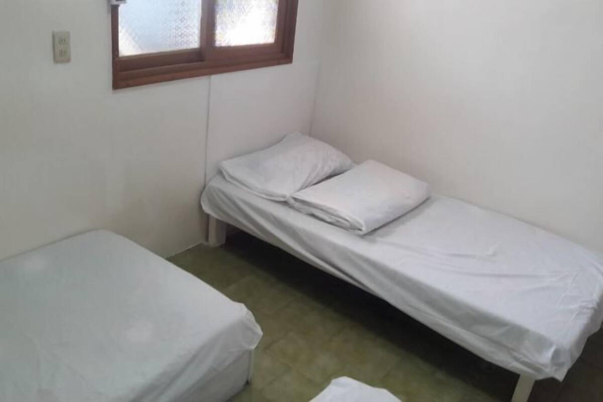 Ein kleines Zimmer mit einem Einzelbett, Fenster, schlichten Wänden und einem Fliesenboden.