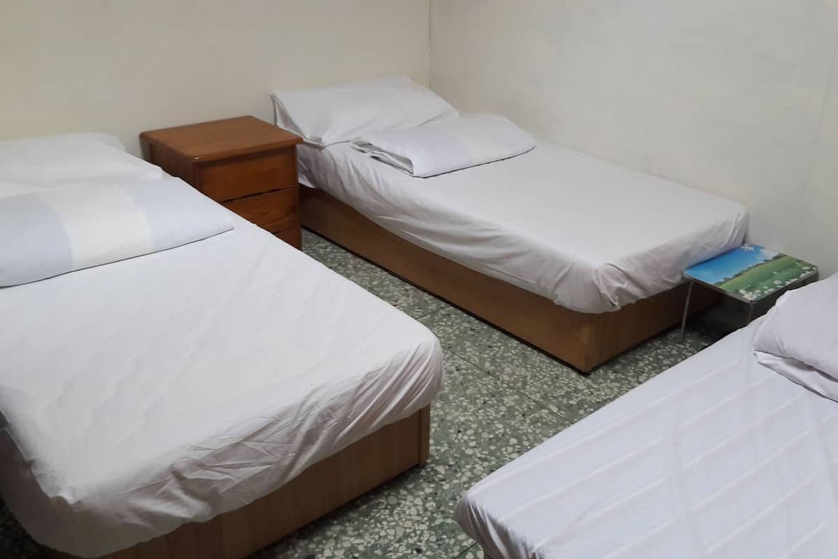 Ein Zimmer mit drei Einzelbetten, jedes mit weißer Bettwäsche und Holz-Kopfteilen. Am linken Rand des Zimmers befindet sich ein kleiner Nachttisch mit einer Schublade. Der Boden ist mit einem fleckigen Fliesenmuster versehen.