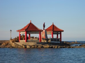 A traditional red-roofed pavilion with a statue on top, situated on a rocky shoreline by the sea.