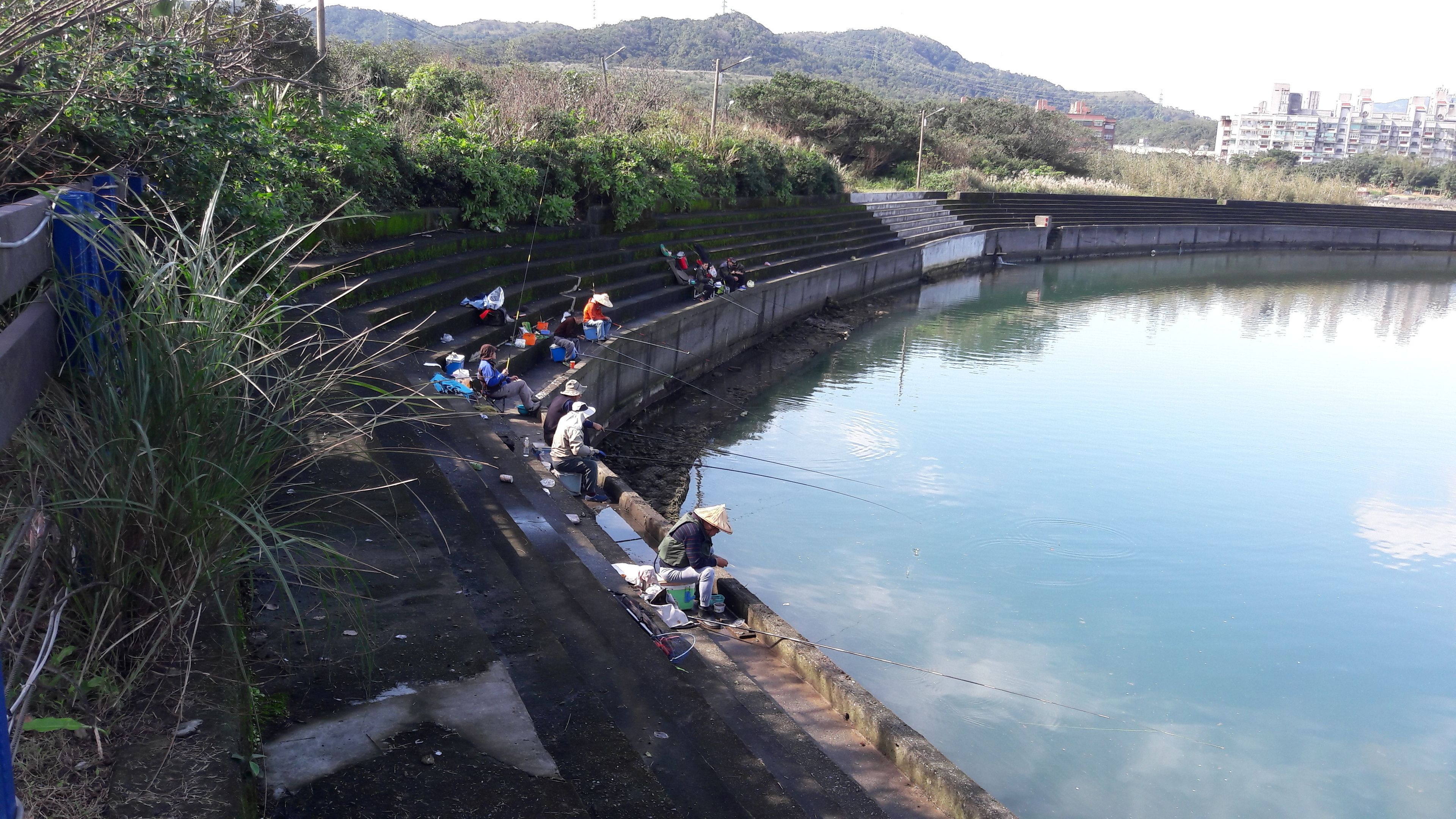 A group of people fishing by a calm body of water with steps leading down to the water's edge.
