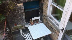 A small outdoor seating area with a white metal table and chairs, a brick wall, and a window. - Stay in Kings Cross - Flat D (London)