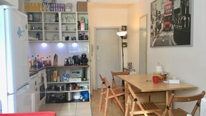 A compact kitchen with white cabinets, a refrigerator, and a dining area with wooden chairs and a table. - Stay in Kings Cross - Flat D (London)