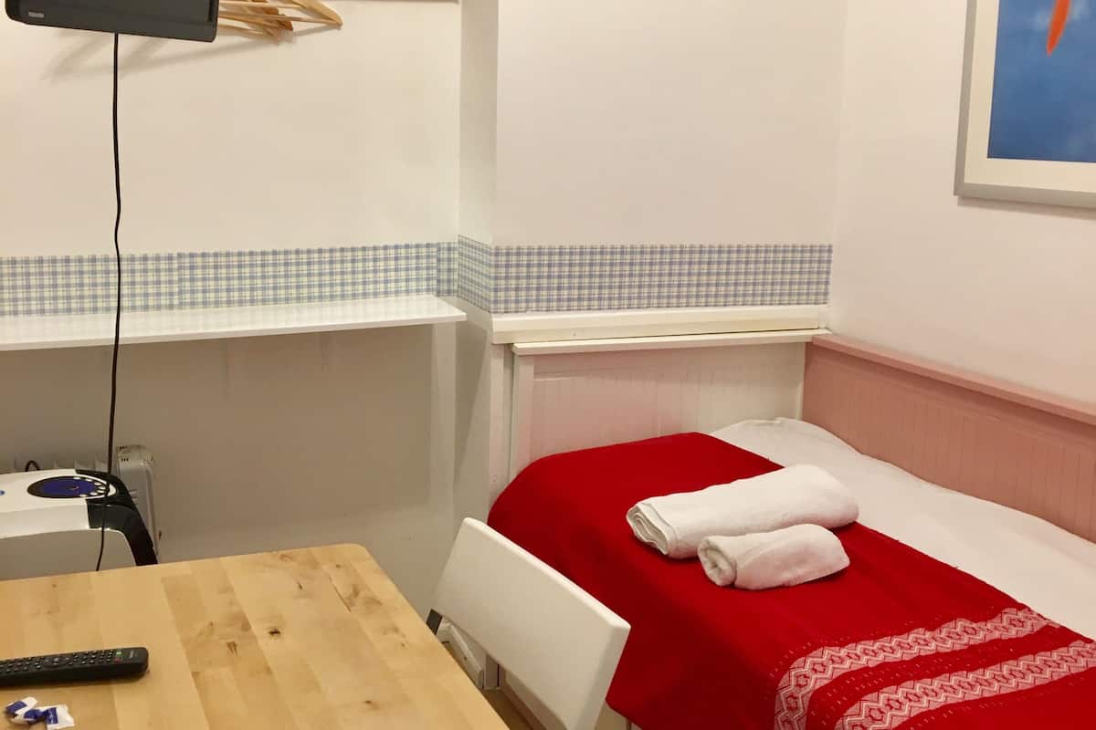 A small, neatly arranged room with a bed, a table, a chair, a TV mounted on the wall, and a shelf with hangers.