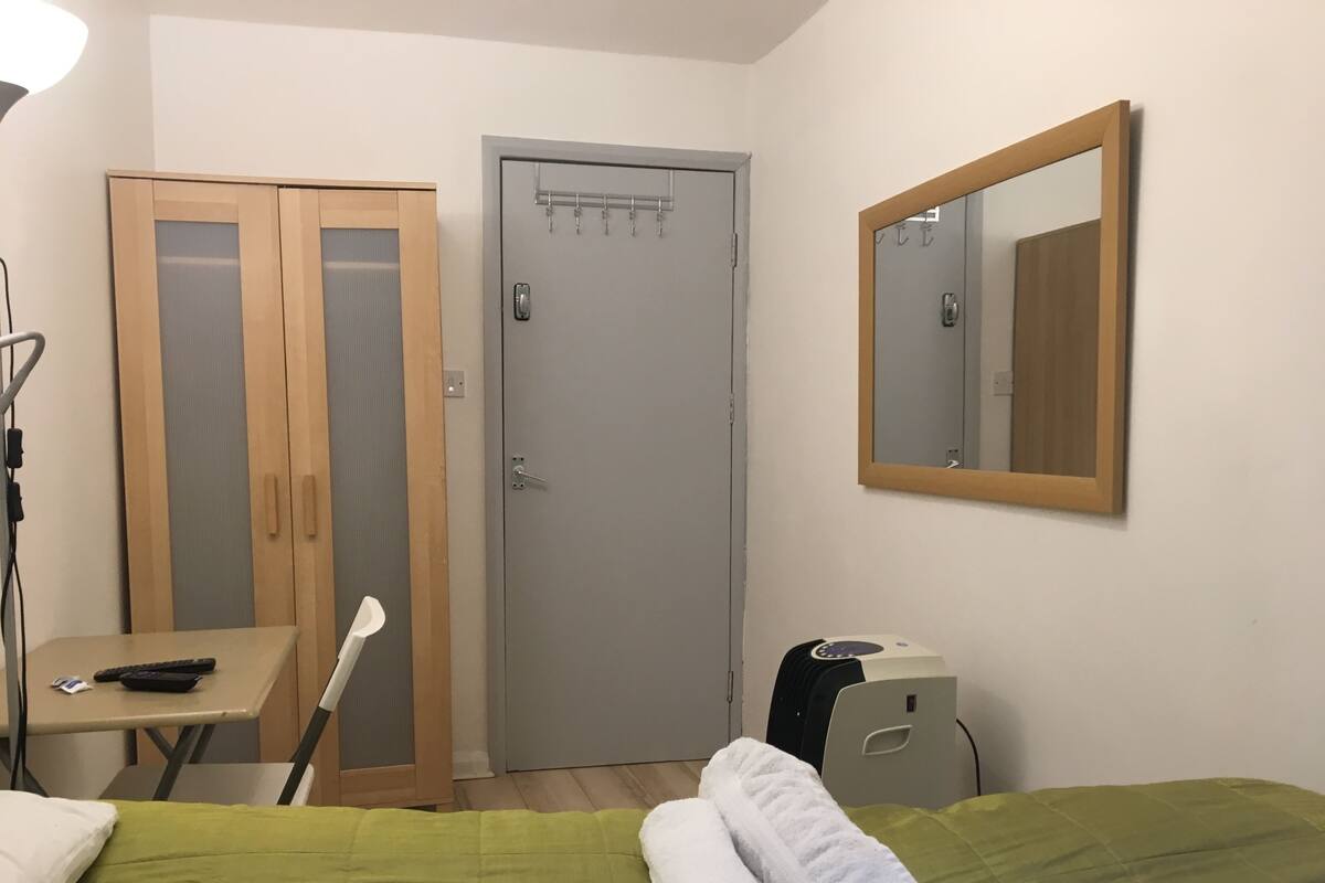 A hotel room with a bed, a desk, a chair, a wardrobe, a mirror, and a small cabinet.