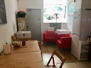 A compact kitchen with a wooden table, chairs, a refrigerator, a washing machine, and a fan.
