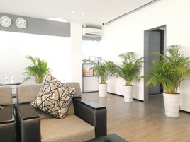 Una sala de estar moderna con un sofá, una mesa de centro y un cojín estampado. Hay varias plantas en macetas y un equipo de aire acondicionado instalado en la pared.