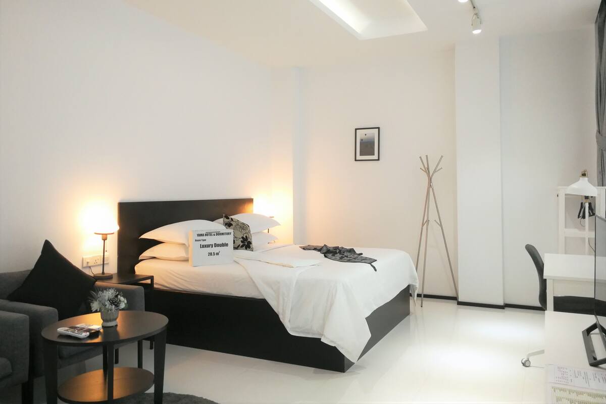 A modern bedroom with a large bed, bedside lamps, a small table with a remote control, and a chair.