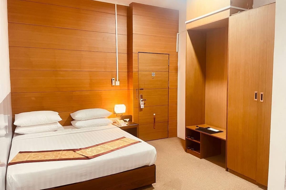A hotel room with a wooden headboard, a bed with white linens, a nightstand with a lamp, and a wardrobe with a mirror.