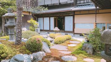 Um jardim japonês tradicional, com lanternas de pedra, um edifício de madeira com portas de correr e um jardim de rochas e musgo bem cuidado.