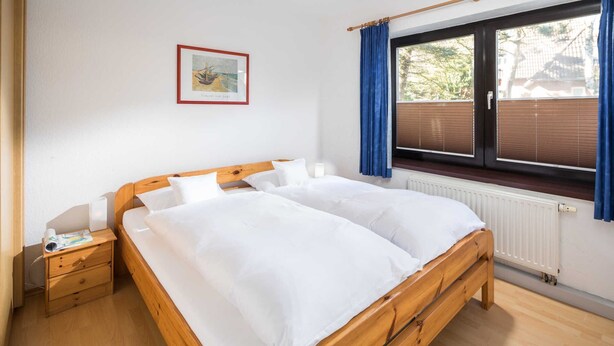 A room with a wooden bed, a nightstand, a window with blinds, and a framed picture on the wall. - 3-room apartment - Logierhaus Birkenhof (Norderney)