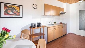 A modern kitchen with wooden cabinets, a sink, and a coffee machine. There is a dining table with chairs and a vase of flowers. - 3-room apartment - Logierhaus Birkenhof (Norderney)