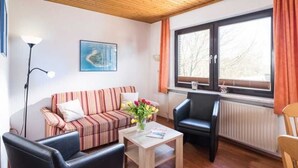 A furnished living room with a striped sofa, a coffee table, a black leather chair, and a window with curtains. - 3-room apartment - Logierhaus Birkenhof (Norderney)