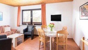 A cozy living room with a sofa, a coffee table, a dining table with chairs, a TV mounted on the wall, and a window with curtains. - 3-room apartment - Logierhaus Birkenhof (Norderney)