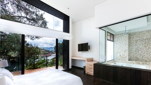 A modern bedroom with a large bed, a glass-enclosed balcony, a wooden desk, and a bathroom with a freestanding bathtub. - Luxé Cabañas (Guatapé)