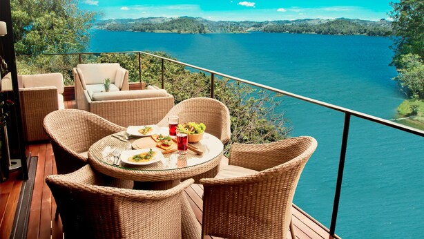 A balcony with wicker furniture and a table set for two, overlooking a lake. - Luxé Cabañas (Guatapé)