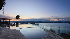 A modern infinity pool with a glass railing, a lounge area with sun loungers, and a view of the water and distant mountains. - Luxé Cabañas (Guatapé)