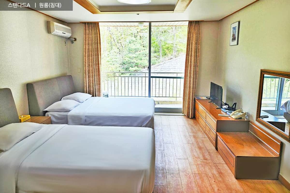 A hotel room with two beds, a desk with a telephone, a television, and a balcony with curtains.