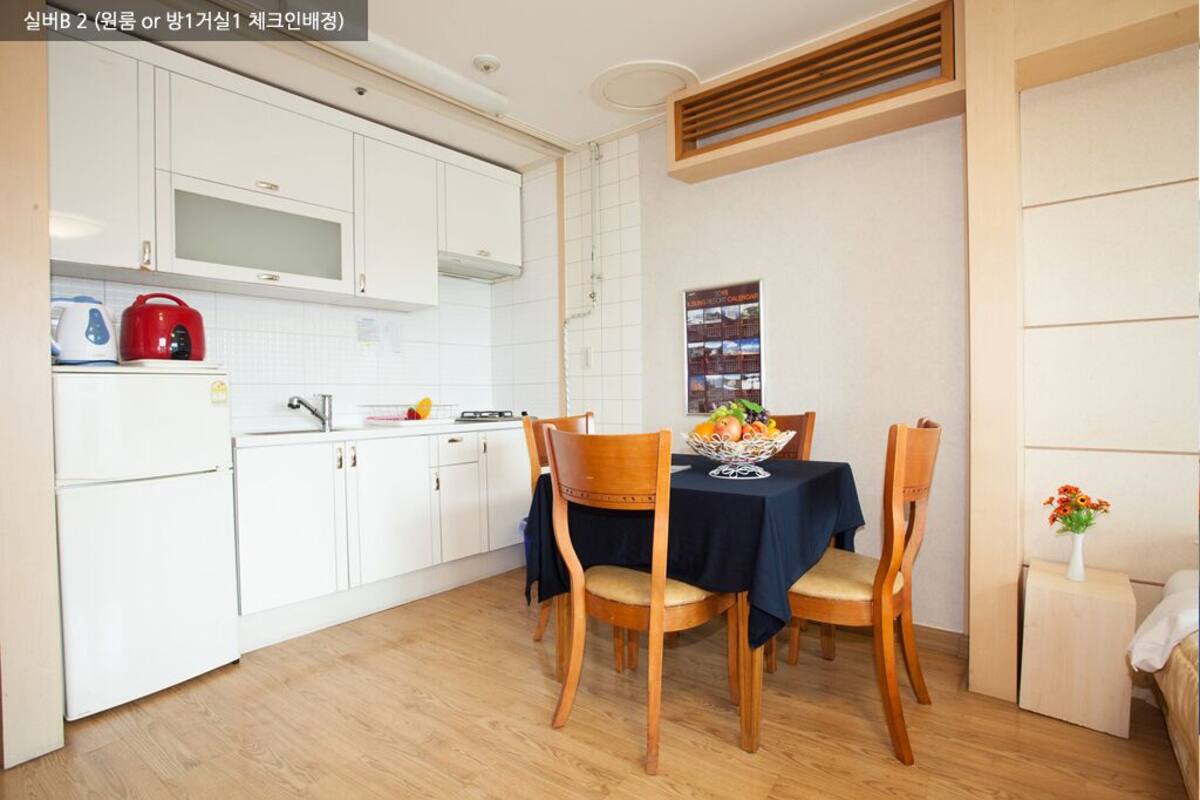 A compact kitchen with white cabinets, a microwave, and a red kettle. There is a dining table with two chairs and a fruit bowl.