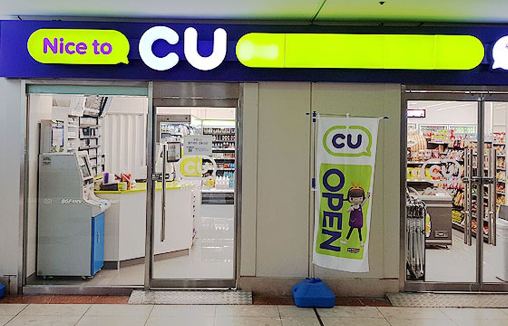 Un magasin CU avec une enseigne « CU OPEN », des portes vitrées et un intérieur visible avec des étagères et des produits exposés.