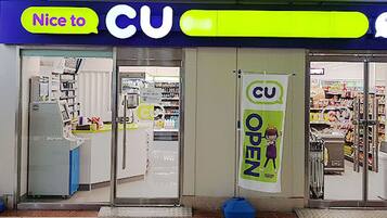 Un magasin CU avec une enseigne « CU OPEN », des portes vitrées et un intérieur visible avec des étagères et des produits exposés.