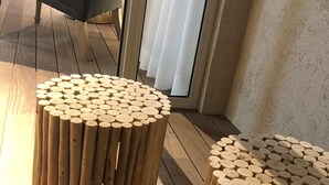 Dos taburetes redondos de madera con detalles recortados, un sofá gris y una mesa de metal con sillas en una habitación con piso de madera y vistas al exterior a través de una ventana con cortinas blancas.