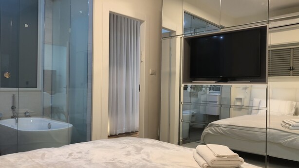 Una habitación de hotel moderna con una cama grande, televisión, baño con paredes de vidrio y un espejo que refleja la habitación.