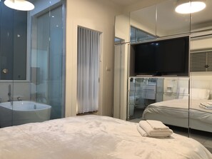 Ett modernt hotellrum med en stor säng, en tv, ett badrum med glasväggar och en spegel som reflekterar rummet.