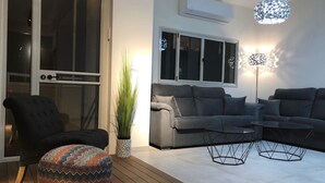 Una sala de estar moderna con un sofá gris, un sillón negro y un reposapiés colorido. Hay una planta en maceta y una lámpara de pie.