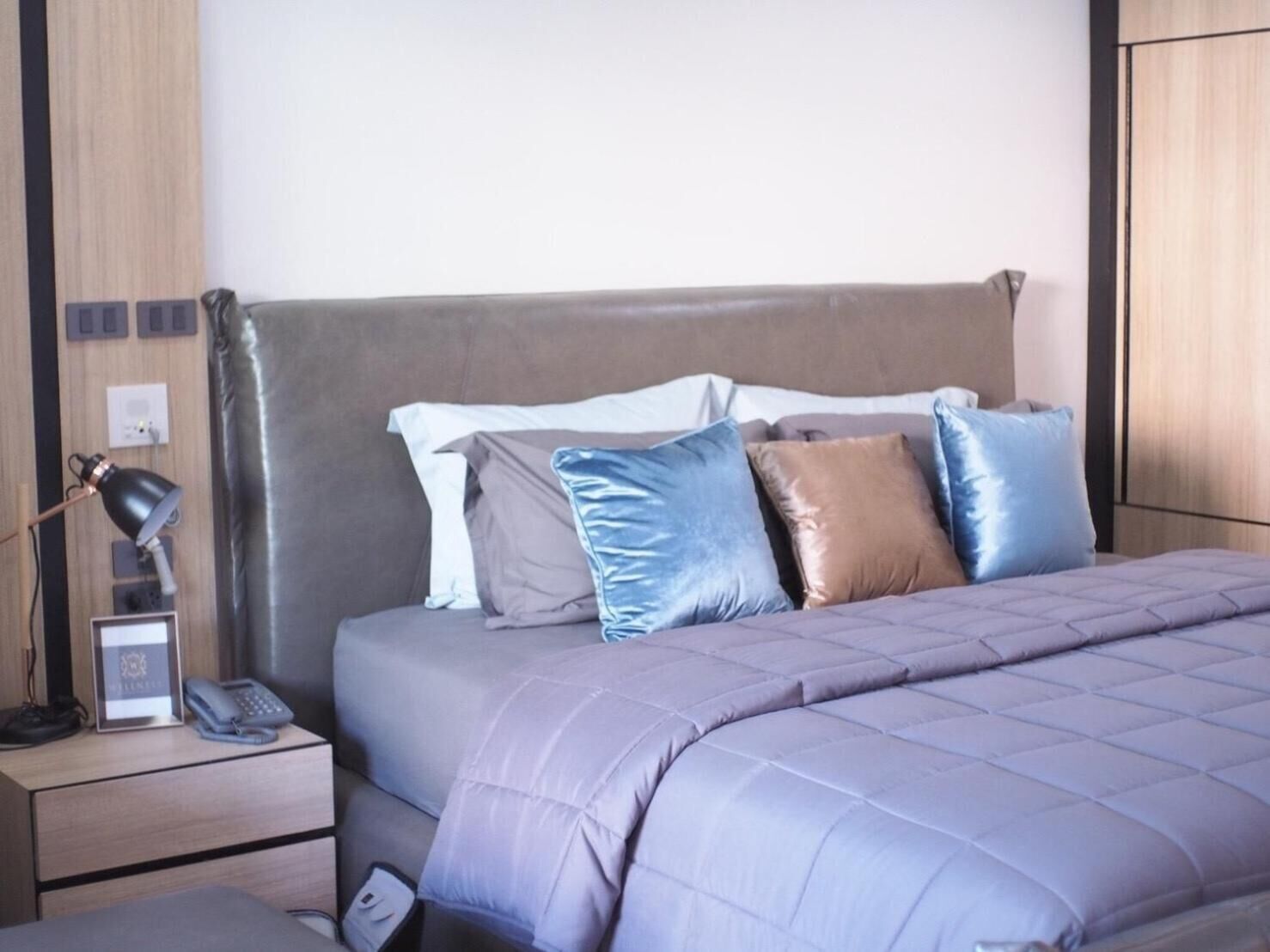 A neatly made bed with a grey headboard, a bedside table with a lamp and a phone, and a wall socket.