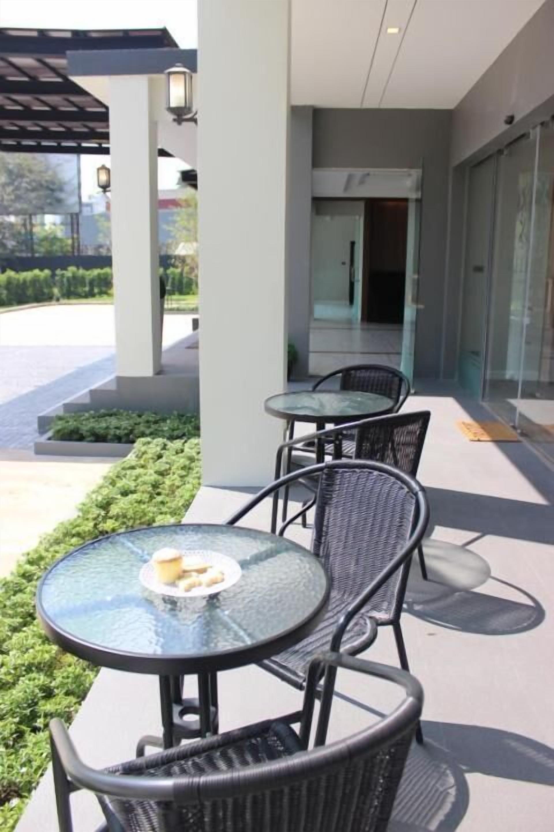 An outdoor seating area with glass tables and wicker chairs, a covered walkway, and a building entrance.