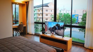 A modern hotel room with a large bed, a desk with a chair, a television on a wooden stand, and a view of the city through large windows.