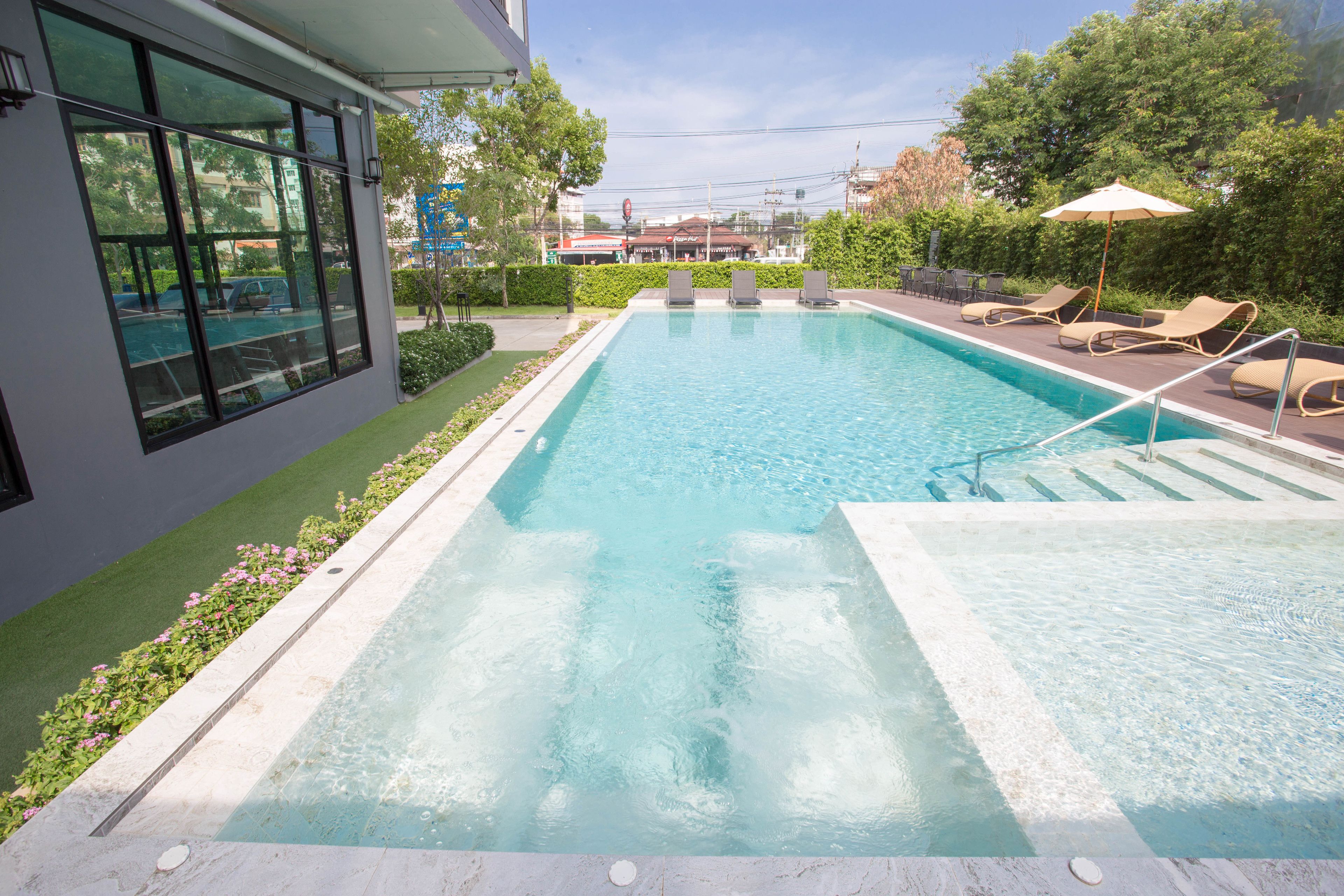 A rectangular swimming pool with a clear blue water, surrounded by a paved deck. There are lounge chairs and a small umbrella on the right side.