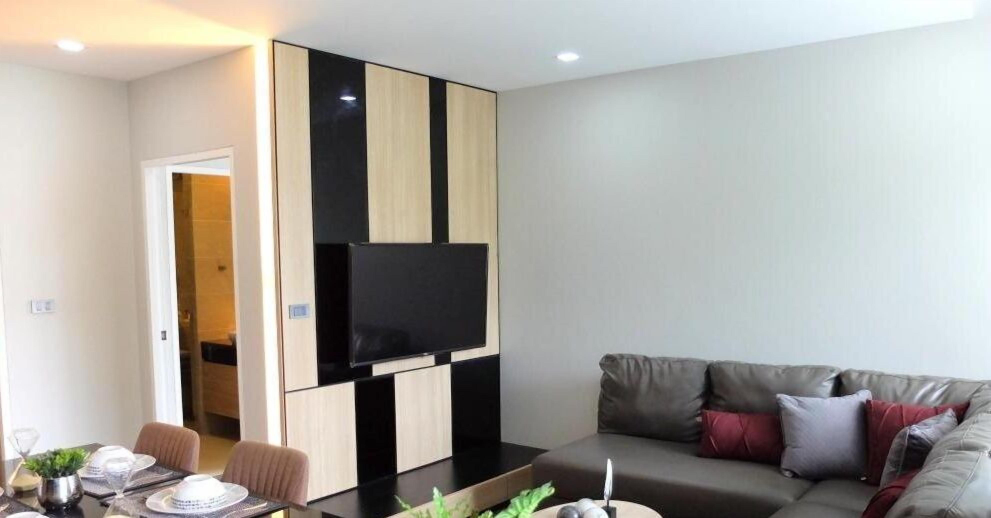 A modern living room with a flat-screen TV mounted on a black and beige panel, a grey sofa with red cushions, and a dining area with a table set for two.