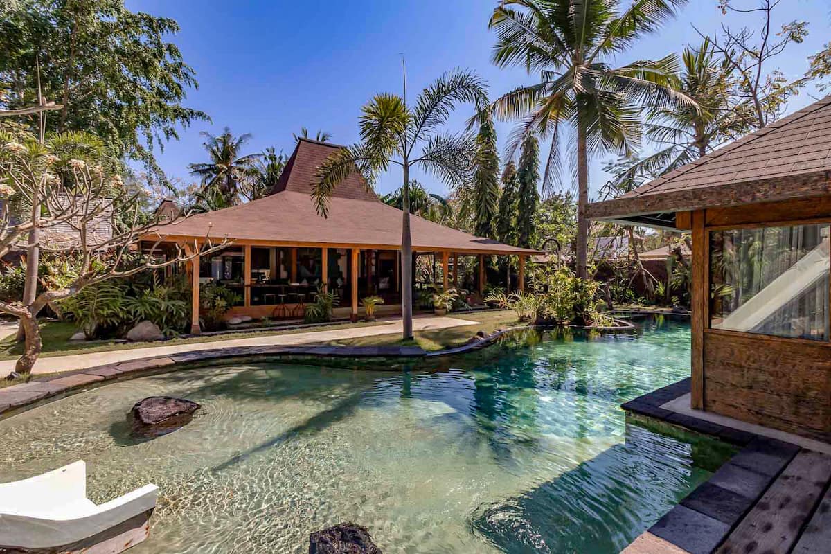 A clear swimming pool with a wooden deck, surrounded by tropical vegetation and a covered outdoor seating area.