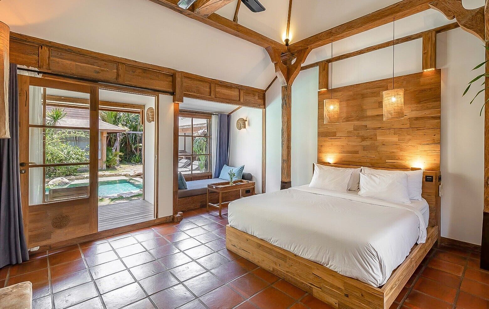 Una camera da letto con un letto grande, travi a cassetto in legno, una porta scorrevole in vetro che dà su una zona piscina e un tavolino con una sedia.