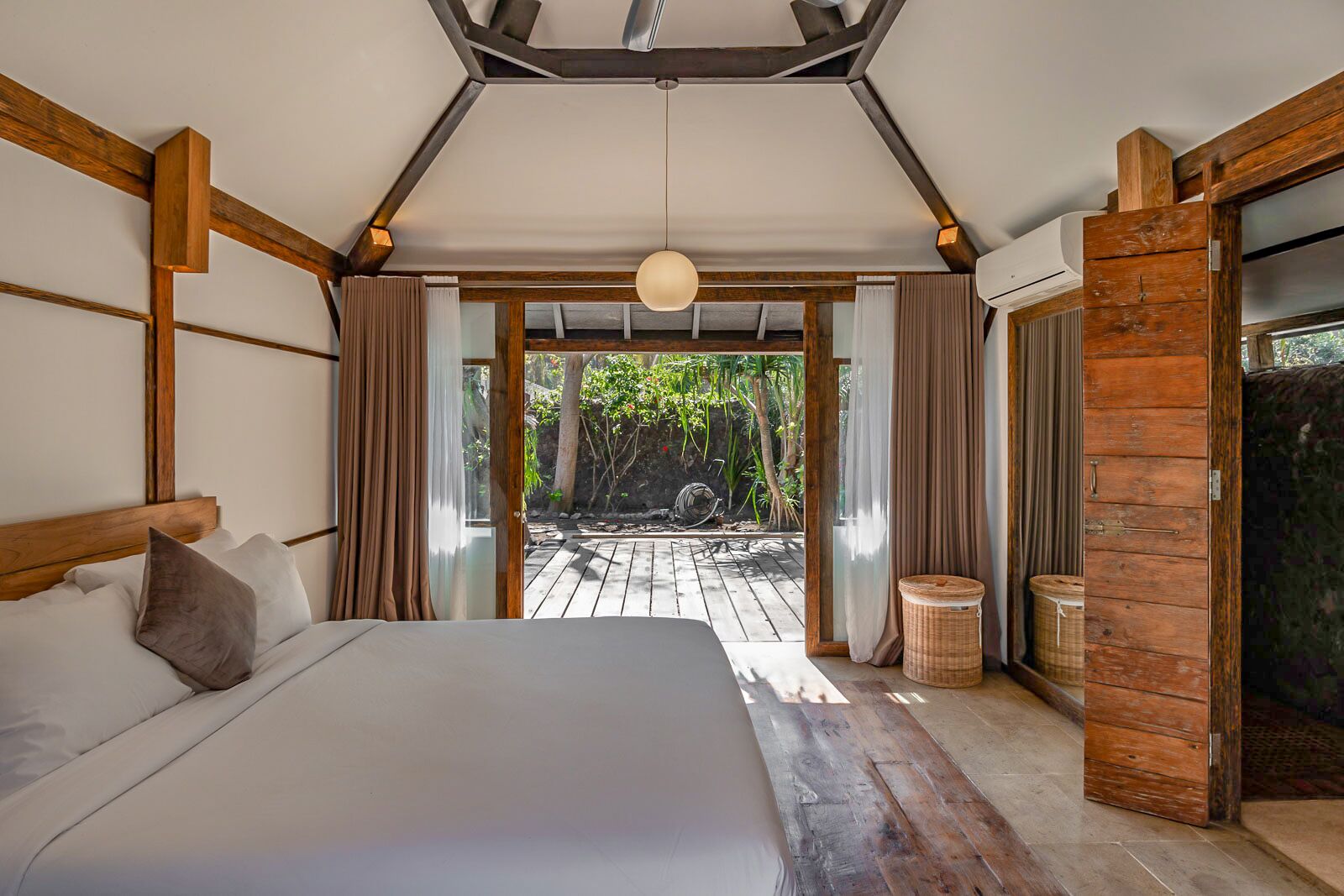 A bedroom with a large bed, wooden beams, a ceiling light, and a view of an outdoor garden.
